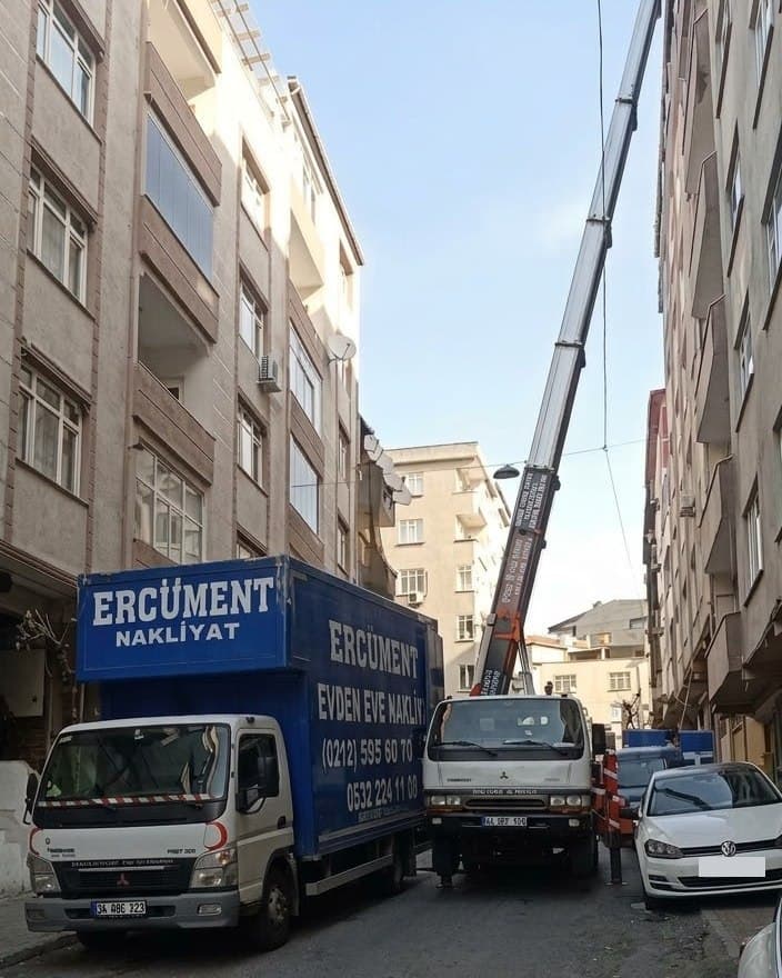 Dar bir sokakta Ercüment Nakliyat kamyonu ve vinçli taşıma aracı birlikte çalışıyor